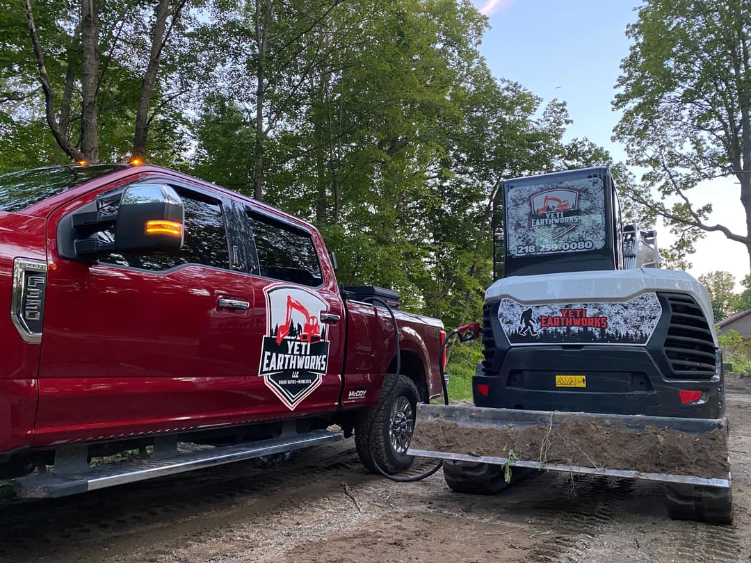 Red truck and mini excavator with Yeti Earthworks branding in a forested area.