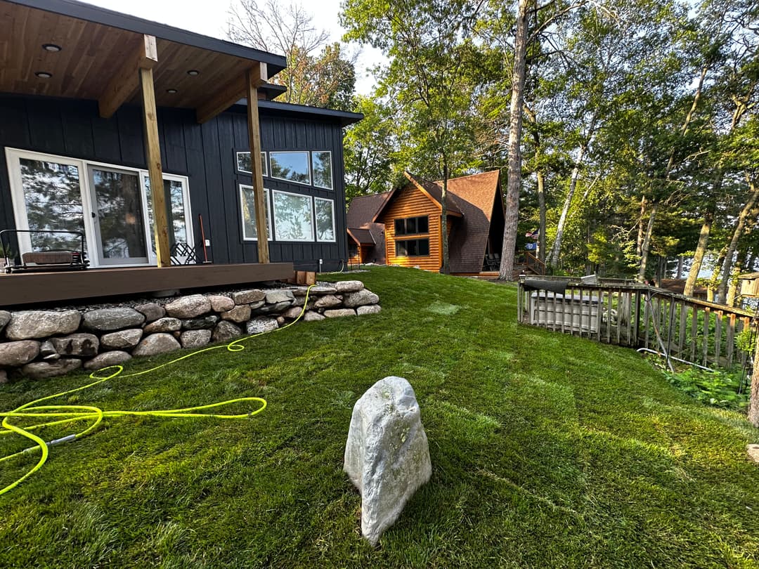 Modern lakeside home with landscaped yard, stone wall, and two rustic cabins in the background.