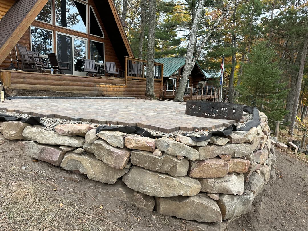 Stone patio with fire pit in front of a log cabin surrounded by trees. Outdoor seating area.