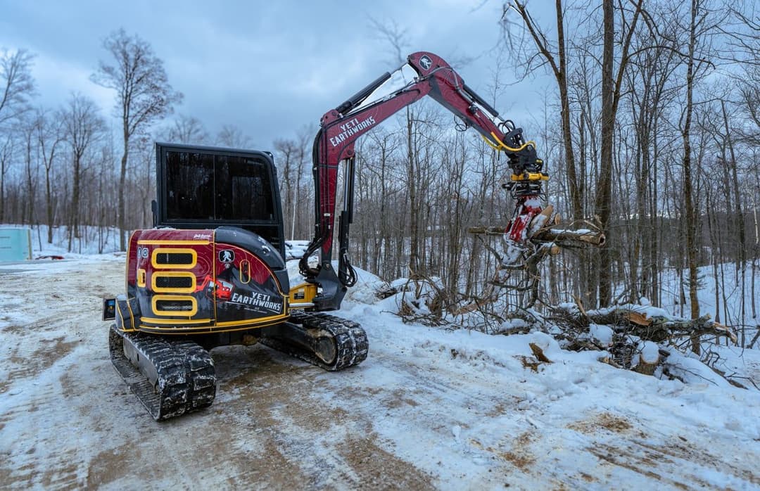 Excavator with a grapple attachment clearing fallen trees in a snowy forest.