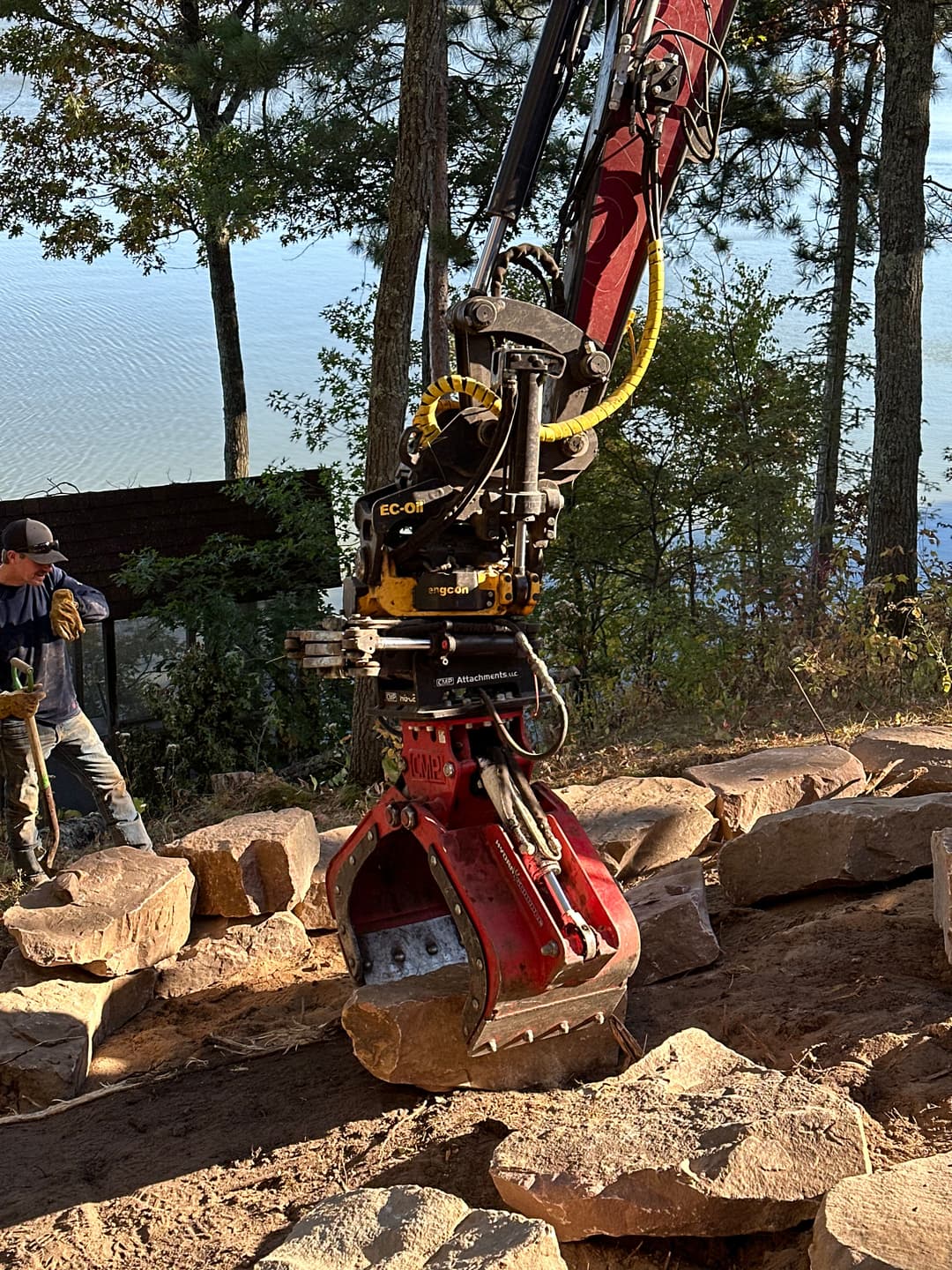 Excavator lifting large rocks near water, with worker in background preparing the site.