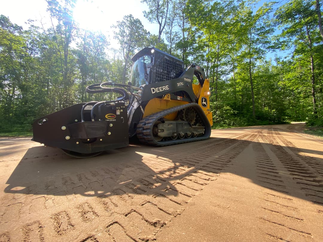 Deere 310SG skid steer loader on a forest road, showcasing tread marks in sunlight.