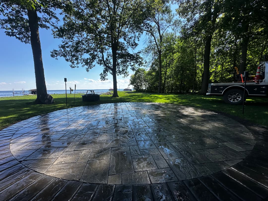 Circular stone patio on a lakeside property with trees and a distant boat. Ideal for outdoor gatherings.