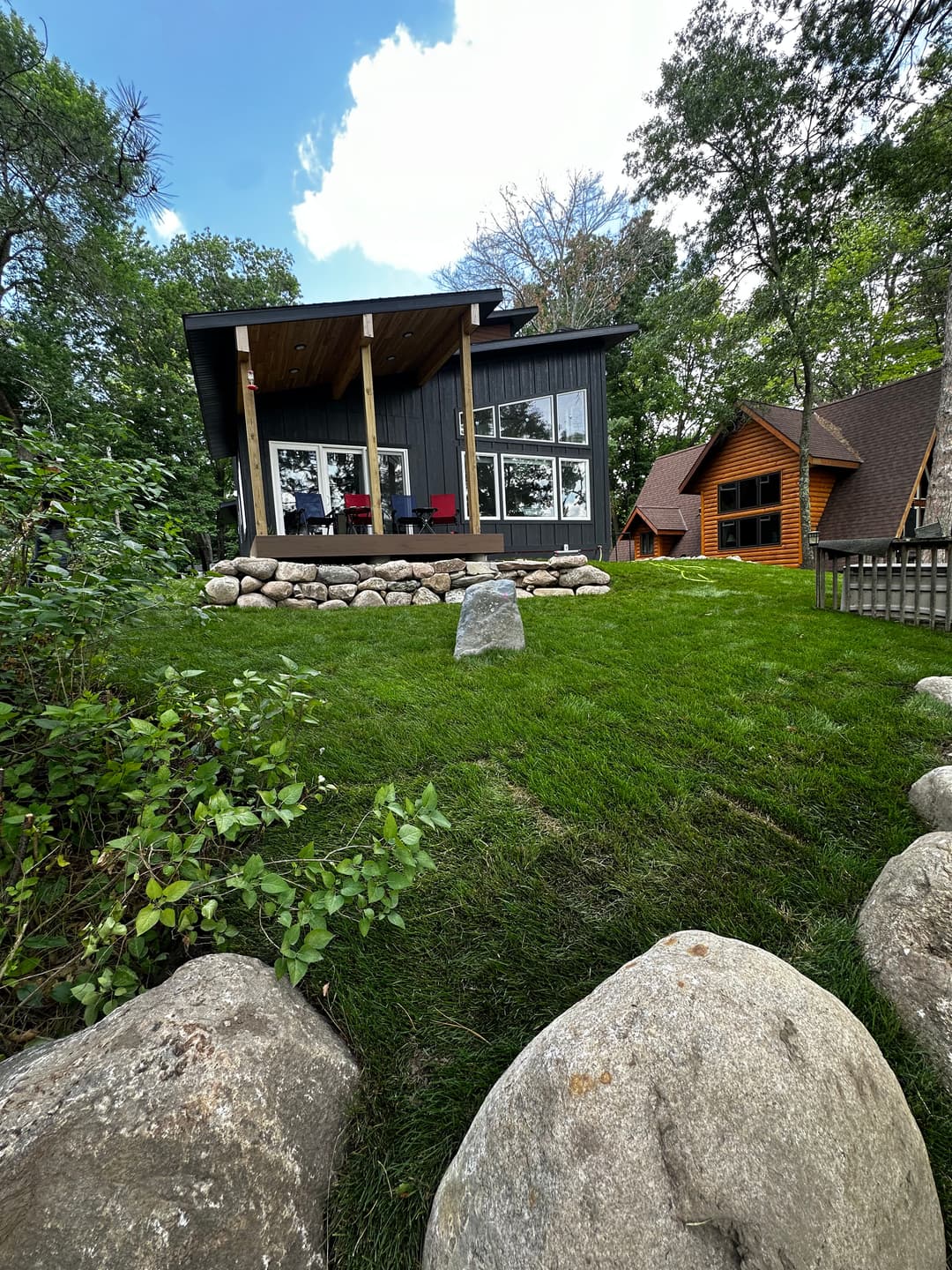Modern cabin with stone landscaping, surrounded by trees and grassy areas on a sunny day.