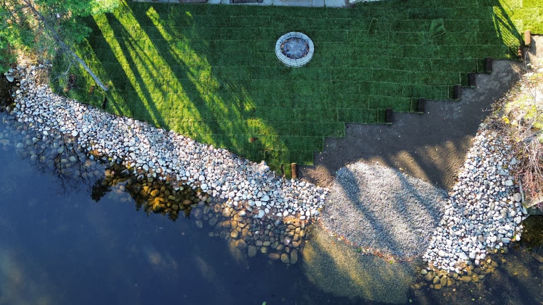 Aerial view of a landscaped lakeshore with grass, stones, and a fire pit.