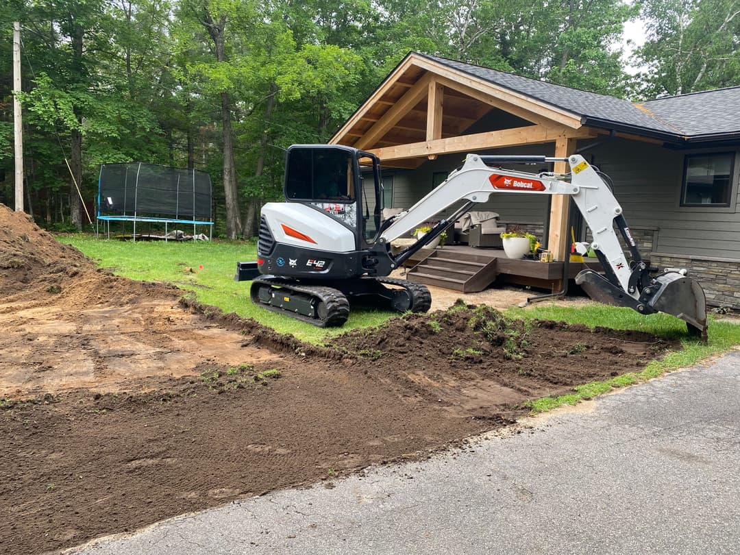 Mini excavator working on landscaping in front of a house with a wooden porch and trampoline.